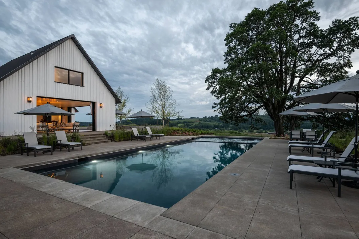 A modern barn-style house is set behind a serene swimming pool, featuring lounge chairs and umbrellas for outdoor relaxation. The area is bathed in natural light, with space for swimming, sunbathing, and casual outdoor meals.