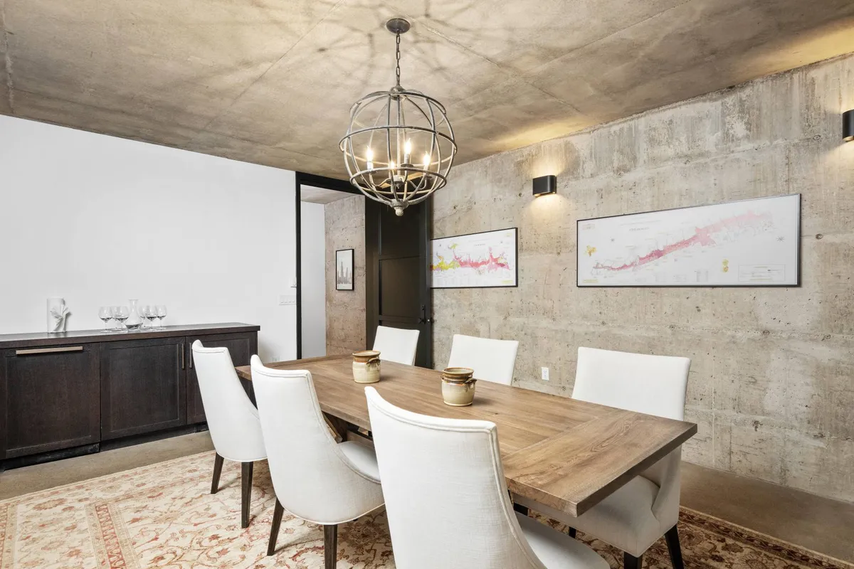 The image features a stylish dining table set beneath a chandelier, with concrete walls and artwork adding to the modern, atmospheric feel. The mood is warm and welcoming, perfect for hosting celebratory toasts or intimate wine tastings.
