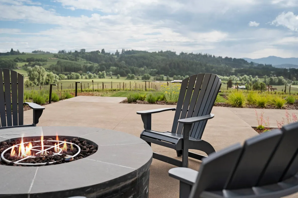 A peaceful exterior scene featuring a cozy fire pit surrounded by Adirondack chairs, set against rolling hills and a distant tree line. The soft evening light suggests a tranquil, sociable setting for sipping wine and enjoying the landscape.