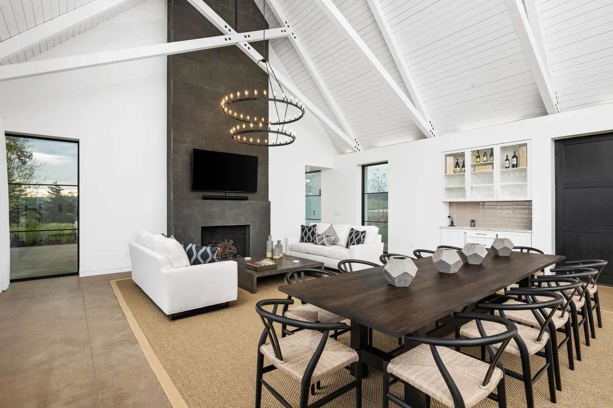 The image showcases the spacious and elegant interior of the Deux Vert Vineyard Estate, with a large dining table perfect for gatherings, a chic chandelier as a focal point, a cozy fireplace beneath a mounted TV, comfortable seating for groups, and expansive windows revealing tranquil valley views.