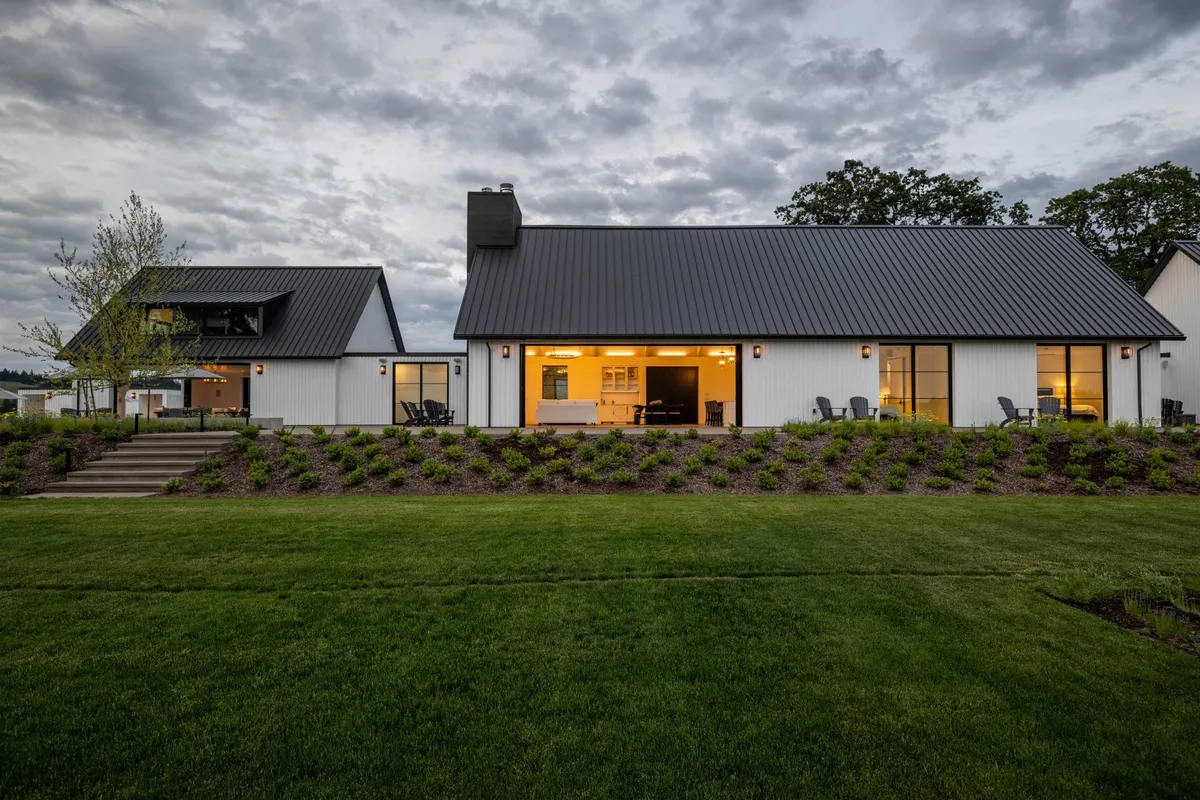 The image showcases the modern house with a sleek metal roof, the warm glow from the windows, and a landscaped yard. The setting illustrates a harmonious connection between the built environment and its natural surroundings, embodying sustainable design.