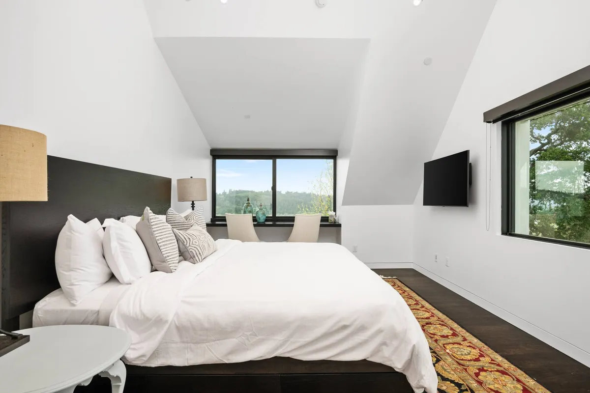 A tranquil king-sized bedroom featuring crisp white linens, a decorative rug, and expansive windows that offer a gorgeous view of the landscape. Natural light fills the space, creating a peaceful retreat ideal for relaxation.