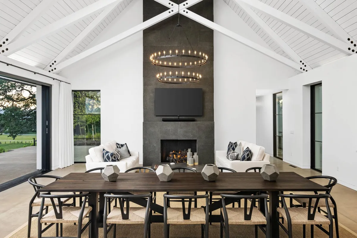 A warm, inviting dining area with a grand table set for a large group, positioned beside a fireplace and beneath a striking chandelier. Floor-to-ceiling windows offer views of the landscape, and the table is perfectly suited for festive gatherings or intimate dinners.