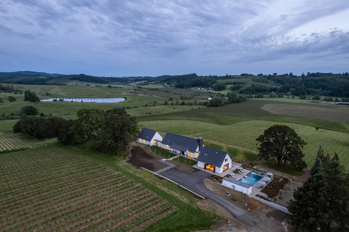 A wide exterior view of a modern farmhouse surrounded by a lush vineyard and rolling hills. The estate features a sparkling swimming pool and ample outdoor space, with neat rows of grapevines extending into the distance under an open sky. The landscape invites visitors to explore both the cultivated vineyard and the tranquil countryside setting.