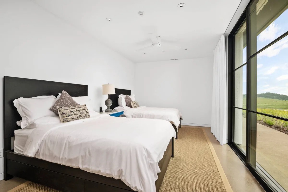A serene bedroom with two beds, large windows bringing in natural light, a lamp, and soft decorative pillows. The layout offers both privacy and togetherness, ideal for families or guests traveling together.