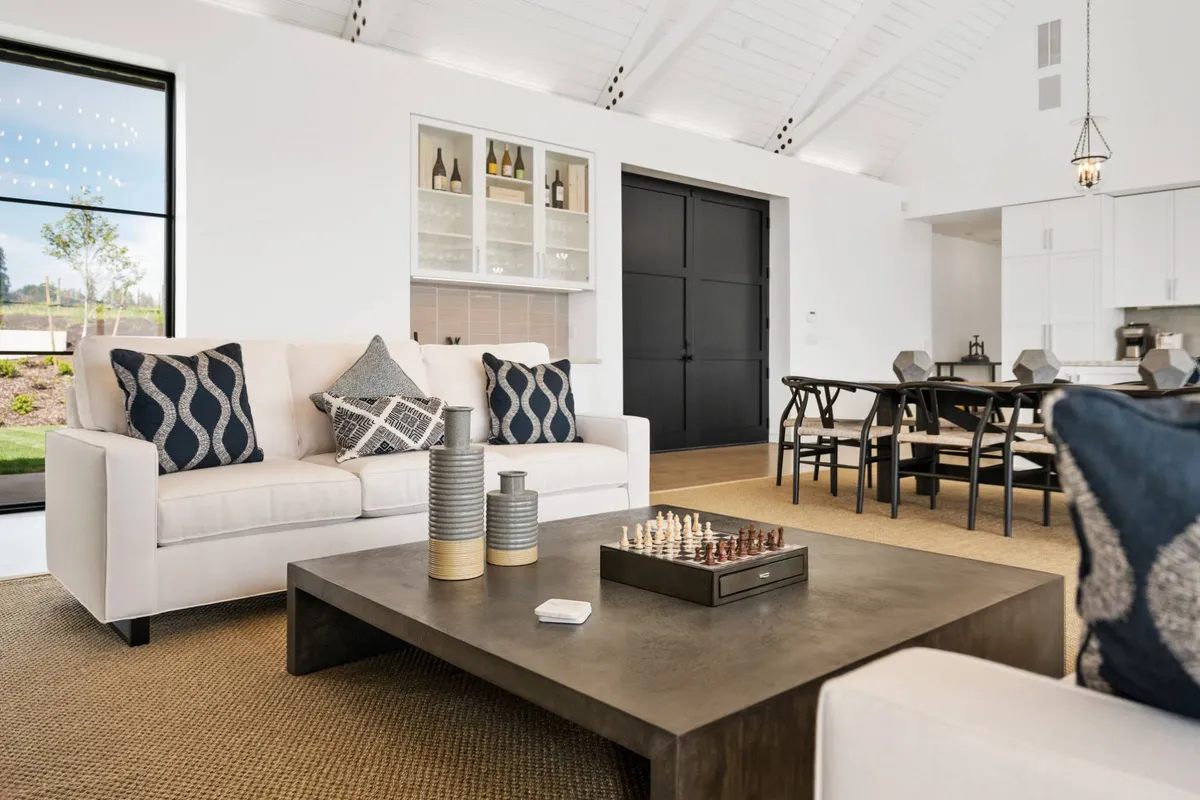 A welcoming interior room featuring a chessboard on a coffee table, a comfortable sofa, and a dining table set up for both socializing and leisure activities. The large windows suggest abundant natural light and a flexible space ideal for gatherings and games.