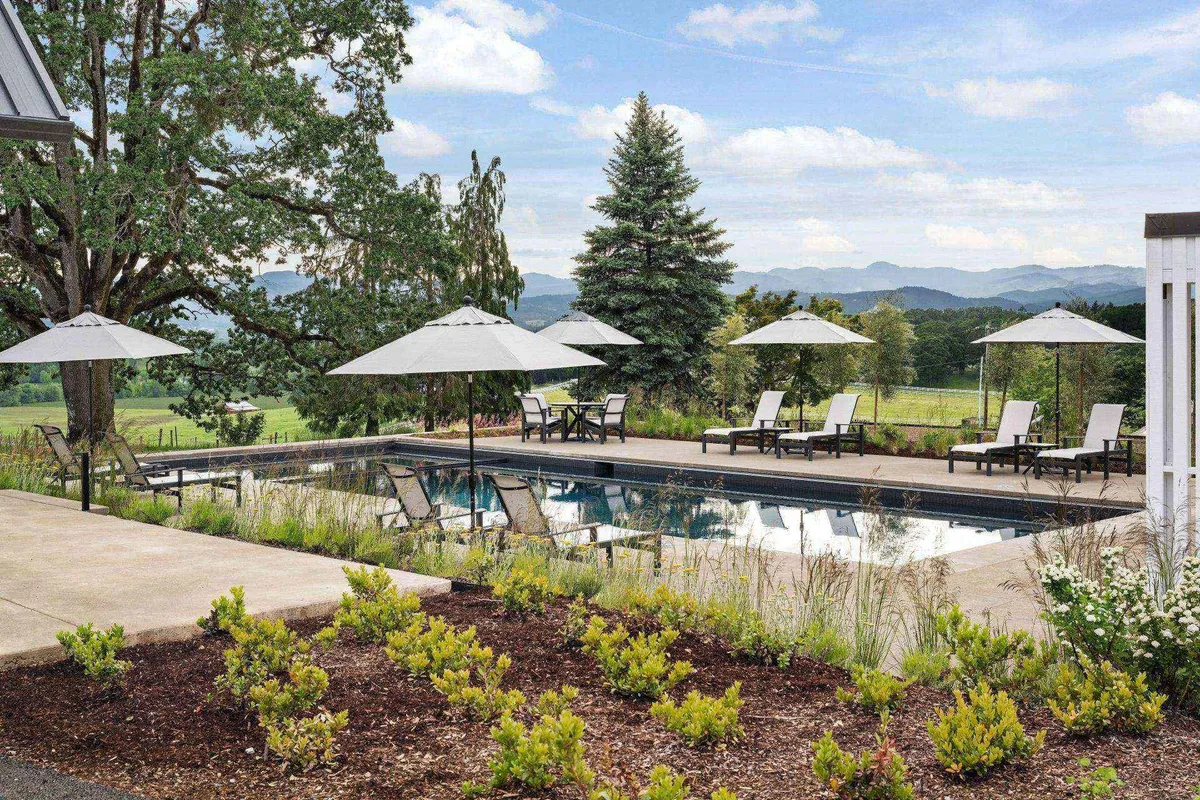 A tranquil swimming pool is surrounded by lounge chairs and umbrellas, set within a landscaped garden. Distant mountains frame the backdrop, while the pool area suggests an inviting place for both energetic swims and quiet relaxation. The scene suggests morning swims, birdwatching opportunities, and a peaceful ambience for wellness activities.