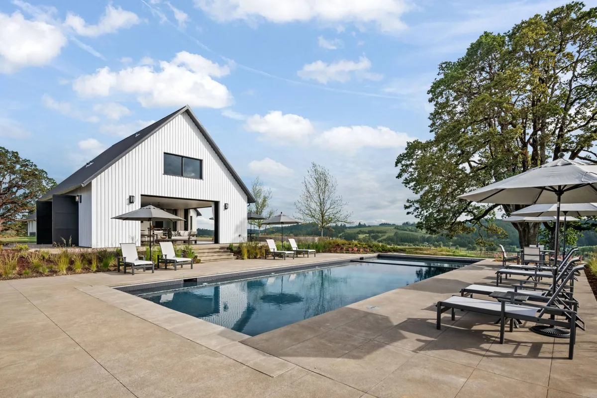 A tranquil exterior scene featuring a modern barn-style house adjacent to a sparkling swimming pool, surrounded by a spacious patio area, lush landscaping, and mature trees. Lounge chairs and patio seating invite group gatherings, with plenty of space for sunbathing, outdoor dining, and social events. The setting hints at both daytime relaxation and lively evening get-togethers.