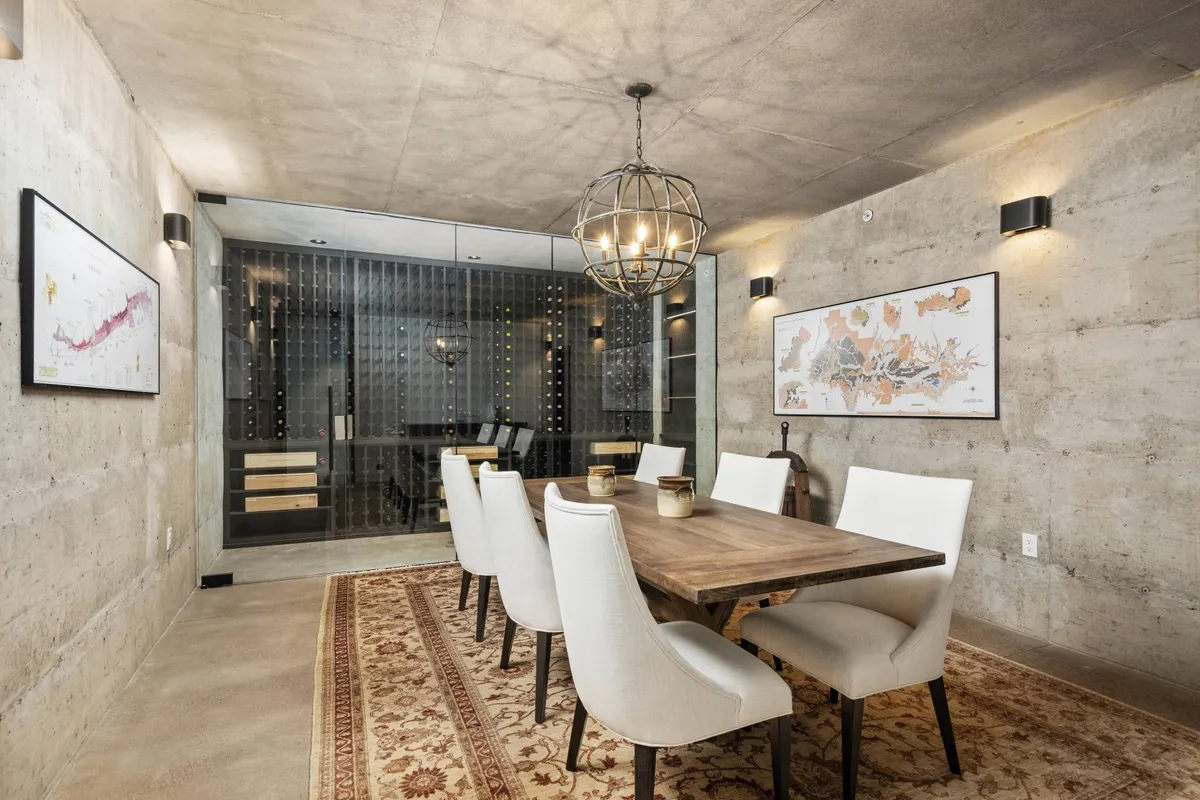 This image showcases a large wooden dining table surrounded by elegant chairs, with a chandelier above and a wine storage area visible in the background. The space exudes sophistication and warmth, suggesting an intimate setting for wine tastings or special gatherings.